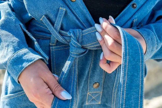 A close-up of hands carefully examining the fabric and stitching of a vintage denim jacket on a hanger in a dimly lit, cozy thrift store.