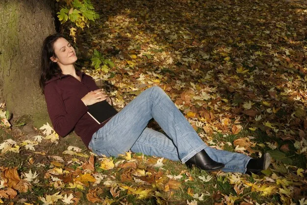A person wearing high-waisted flared jeans in a medium wash, paired with a fitted knit top and block-heel boots, against a backdrop of autumn leaves.