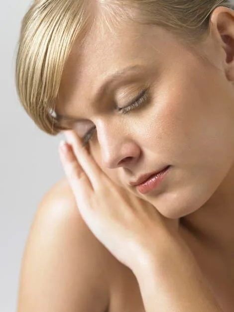 Close-up of a person gently touching their cheek, showing slightly flaky, tight skin, indicating dryness. The lighting is soft to emphasize skin texture.