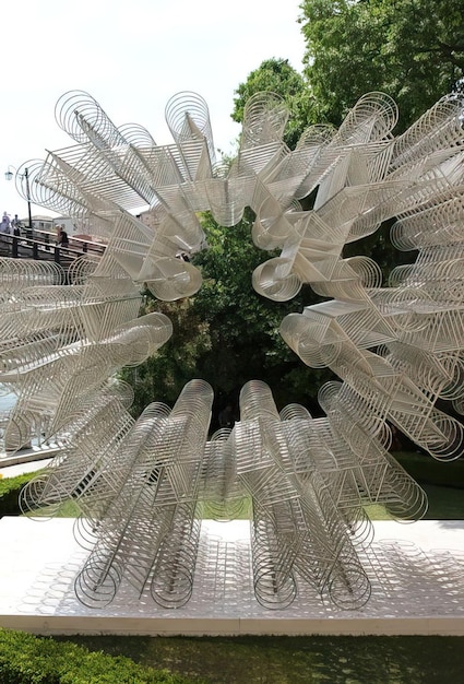 A large, abstract metal sculpture stands in the center of a newly developed urban park. Families are picnicking nearby, and children are playing around the sculpture. The background features modern buildings and a clear sky, showcasing the integration of art into the urban landscape.