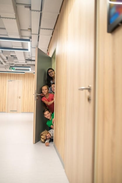 A brightly lit school hallway with students participating in a lockdown drill. One teacher is closing a door while the students crouch quietly against the wall