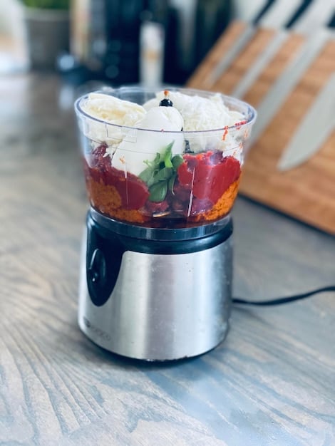 A compact, sleek mini food processor in a modern kitchen setting, actively chopping fresh herbs. The kitchen counter is minimalist, with natural light streaming in, highlighting the device's efficiency and small footprint. A timer on the oven in the background shows just a few seconds, emphasizing speed.
