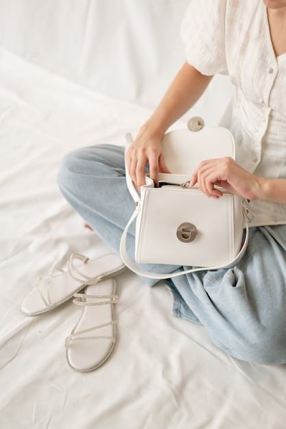 A close-up shot of a minimalist, structured handbag in a neutral beige tone on a clean, light-colored background. The lighting is soft, highlighting the bag's clean lines and simple hardware.